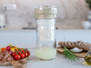 Bodegón mediterráneo con el fermentador de queso de Kéfir Kefirko como pieza central, junto a pan, tomates y aceite de oliva.